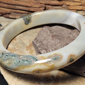 May include: A smooth, polished bangle bracelet crafted from translucent white agate stone. The circular band features natural inclusions of mossy green, grey, and earthy brown mineral patterns embedded within the semi-transparent material.