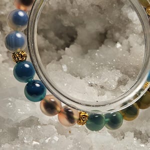 May include: A colourful beaded bracelet with round gemstones in shades of blue, green, pink, and yellow. Gold-coloured decorative elements separate the beads. The bracelet is displayed on a white, crystalline surface.