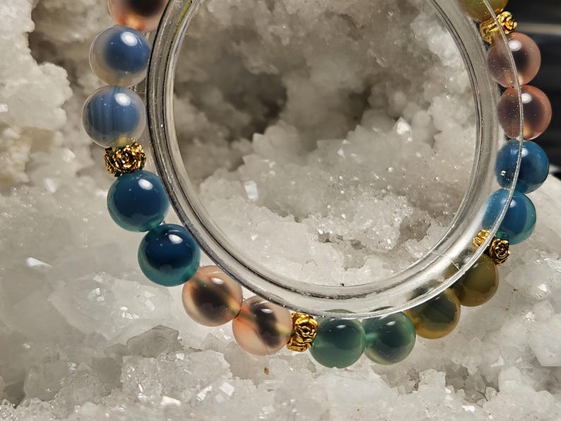 May include: A colourful beaded bracelet with round gemstones in shades of blue, green, pink, and yellow. Gold-coloured decorative elements separate the beads. The bracelet is displayed on a white, crystalline surface.