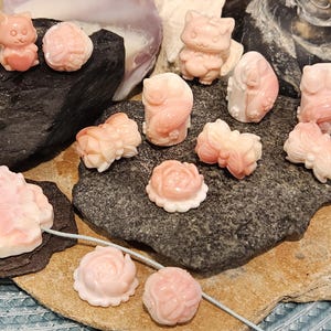 May include: A collection of pink coral carvings, including flowers, animals, and abstract shapes. The carvings are displayed on a dark gray rock surface.