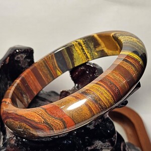 May include: A polished bangle bracelet crafted from tiger's eye stone, featuring a smooth, rounded surface with distinct, layered bands of golden-yellow, deep brown, and reddish-orange hues that create a natural, chatoyant striped pattern.