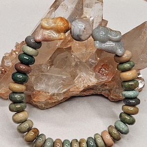 May include: A beaded bracelet featuring a variety of earth-toned stones. The bracelet includes two carved stone rabbit charms and a round, gray and white stone. The beads are in shades of green, brown, and tan.
