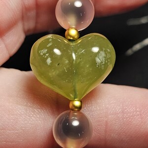 May include: A close-up of a beaded bracelet featuring a heart-shaped green stone, gold accents, and translucent beads in shades of pink and blue. The bracelet is strung together with a delicate cord.