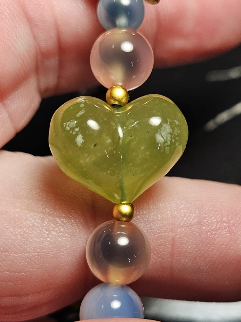 May include: A close-up of a beaded bracelet featuring a heart-shaped green stone, gold accents, and translucent beads in shades of pink and blue. The bracelet is strung together with a delicate cord.