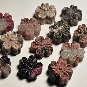 May include: A collection of carved flower-shaped gemstones in various colors, including pink, gray, and black. The gemstones are arranged on a white surface, showcasing their intricate floral designs. These decorative stones are ideal for jewelry making or crafts.