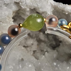 May include: A beaded bracelet with a gold frog charm and beads in blue, pink, and green. The bracelet is strung on a clear, curved support. The beads include a green teardrop shape and several round, translucent beads.