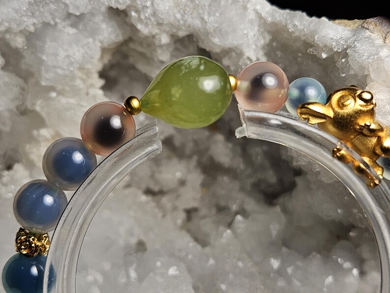 May include: A beaded bracelet with a gold frog charm and beads in blue, pink, and green. The bracelet is strung on a clear, curved support. The beads include a green teardrop shape and several round, translucent beads.