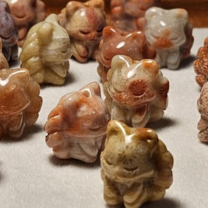 May include: A collection of small, carved cat figurines in various colors, including shades of brown, beige, and gray. The figurines have detailed features and a polished finish, suggesting they are made of stone or a similar material. The cats are arranged on a white surface.