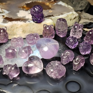 May include: A collection of small, carved amethyst gemstones in various shapes, including foxes, cats, and hearts. The translucent purple stones feature smooth, polished surfaces and intricate details, showcasing the natural colour variations of the quartz.