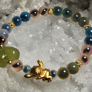May include: A beaded bracelet with a gold-coloured cartoon character charm. The bracelet features blue, pink, and green beads, with gold-coloured spacers. A green heart-shaped bead is also included.