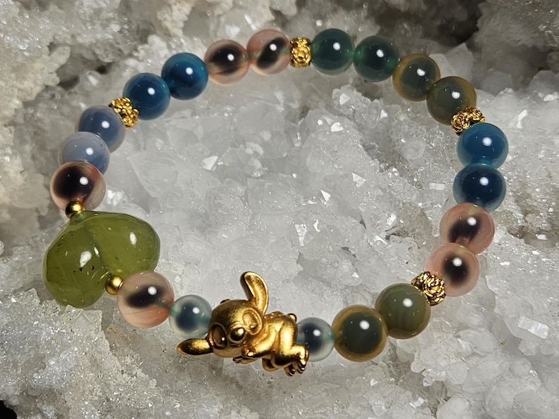 May include: A beaded bracelet with a gold-coloured cartoon character charm. The bracelet features blue, pink, and green beads, with gold-coloured spacers. A green heart-shaped bead is also included.