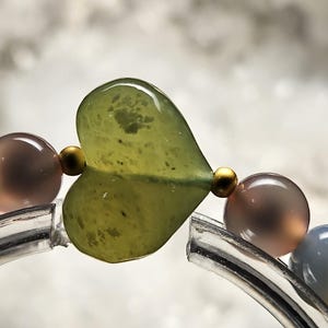 May include: Close-up of a bracelet featuring a large, heart-shaped green bead. The bracelet includes round, translucent beads in shades of grey, brown, and gold. The bracelet is strung on a clear, curved band.