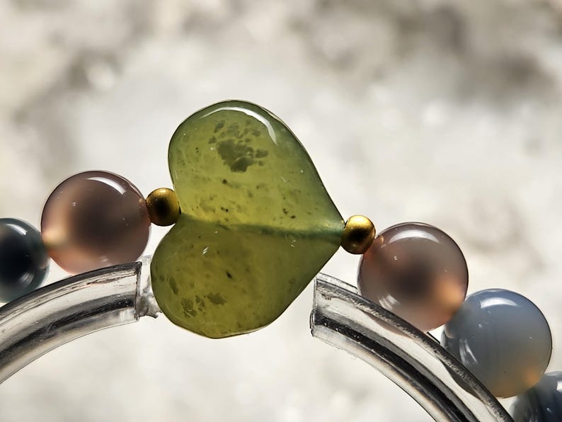 May include: Close-up of a bracelet featuring a large, heart-shaped green bead. The bracelet includes round, translucent beads in shades of grey, brown, and gold. The bracelet is strung on a clear, curved band.