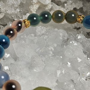 May include: A bracelet with a selection of colourful, round gemstones. The beads are in shades of blue, green, and pink, with gold-coloured decorative elements. The bracelet is set against a backdrop of white crystals.