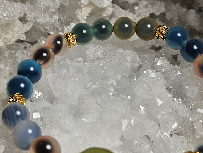 May include: A bracelet with a selection of colourful, round gemstones. The beads are in shades of blue, green, and pink, with gold-coloured decorative elements. The bracelet is set against a backdrop of white crystals.