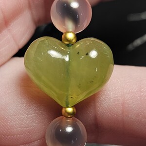 May include: A close-up of a beaded bracelet featuring a heart-shaped green gemstone bead. The bracelet also includes round beads in shades of pink and blue, separated by gold-coloured spacers. The bracelet is strung on a flexible cord.