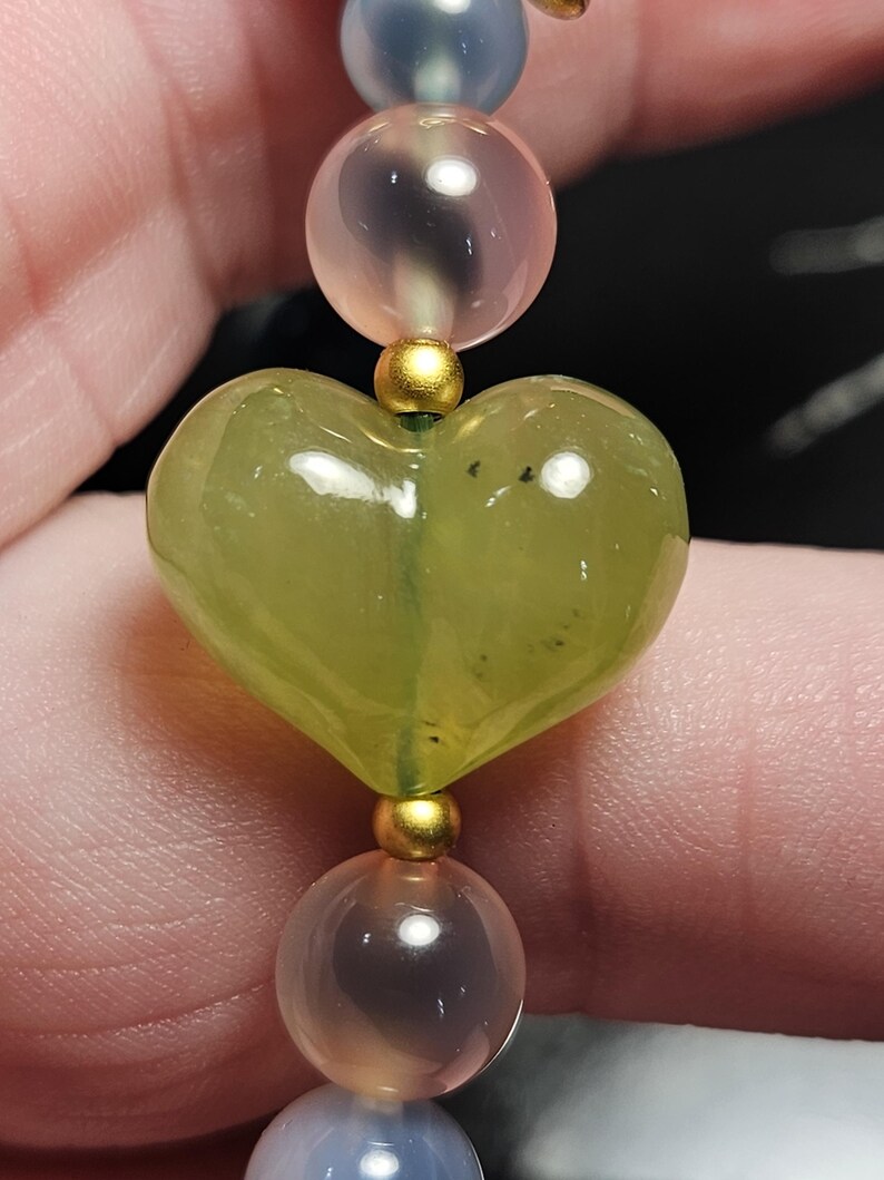 May include: A close-up of a beaded bracelet featuring a heart-shaped green gemstone bead. The bracelet also includes round beads in shades of pink and blue, separated by gold-coloured spacers. The bracelet is strung on a flexible cord.