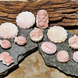 May include: A collection of carved pink and white gemstone figurines, including fish, flowers, and circular designs. The carvings are displayed on a dark gray stone surface with a wooden background.