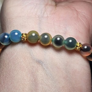 May include: A colourful beaded bracelet with translucent beads in shades of blue, green, yellow, and peach. The beads are separated by small, gold-coloured decorative elements. The bracelet is worn on a wrist.