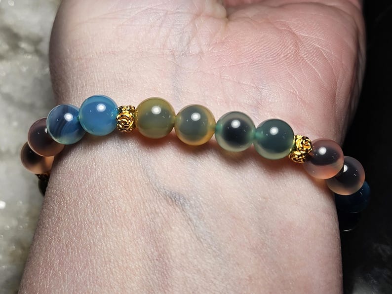 May include: A colourful beaded bracelet with translucent beads in shades of blue, green, yellow, and peach. The beads are separated by small, gold-coloured decorative elements. The bracelet is worn on a wrist.
