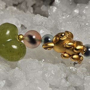May include: A bracelet with a gold koala charm and a green heart-shaped bead. The bracelet also features pink, blue, and green round beads. The background is a white, crystalline surface, resembling quartz or similar mineral formations.
