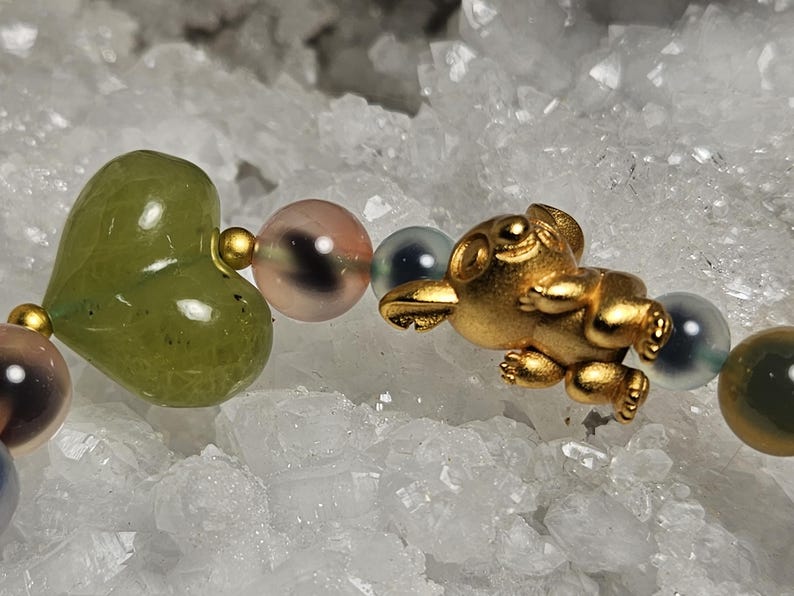 May include: A bracelet with a gold koala charm and a green heart-shaped bead. The bracelet also features pink, blue, and green round beads. The background is a white, crystalline surface, resembling quartz or similar mineral formations.