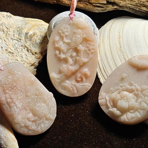 May include: Three light pink pendants with carved designs. One pendant features a dragon, another a flower, and the third an abstract design. Each has a pink cord for hanging.