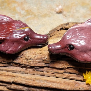 May include: Two red jasper fox carvings, one with a yellow stripe, facing each other on a piece of driftwood.