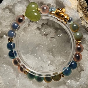 May include: A colourful beaded bracelet with a green heart-shaped charm, gold details, and a gold figurine. The bracelet features blue, pink, and green beads. The bracelet is displayed on a white, textured surface.