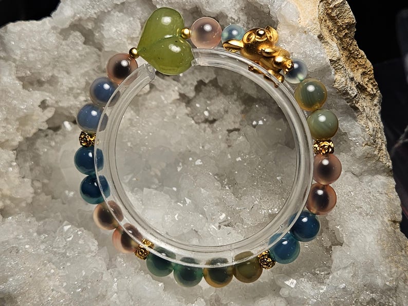 May include: A colourful beaded bracelet with a green heart-shaped charm, gold details, and a gold figurine. The bracelet features blue, pink, and green beads. The bracelet is displayed on a white, textured surface.