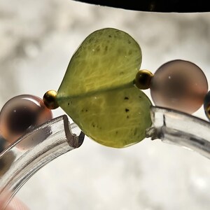 May include: A close-up of a bracelet featuring a heart-shaped, pale green bead. The bracelet also includes round, translucent beads in shades of grey and brown, and small gold-coloured beads. The bracelet is on a clear, curved band.