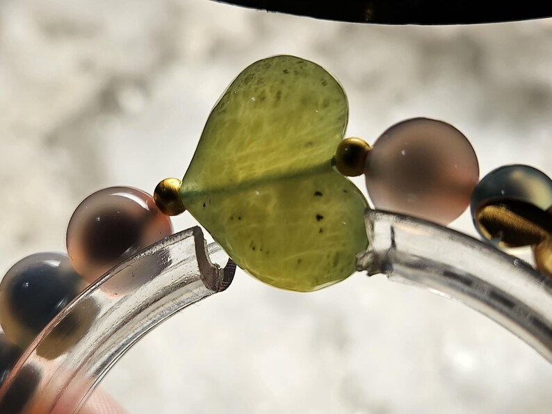 May include: A close-up of a bracelet featuring a heart-shaped, pale green bead. The bracelet also includes round, translucent beads in shades of grey and brown, and small gold-coloured beads. The bracelet is on a clear, curved band.
