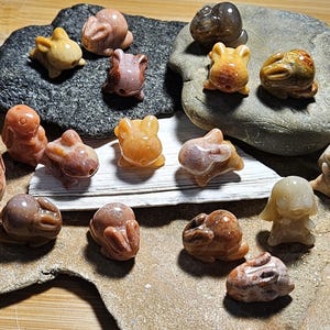 May include: A collection of small, carved stone animal figurines, including rabbits, bears, and other creatures. The figurines are in shades of brown, tan, and orange, and are displayed on a stone surface.
