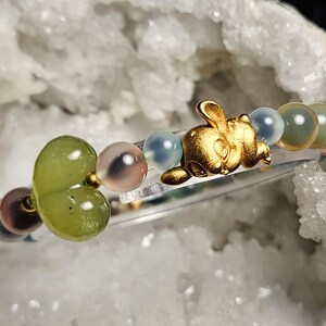May include: A bracelet with a gold-coloured rabbit charm and assorted gemstone beads in green, pink, blue, and brown. The bracelet is strung on a clear elastic cord. The beads are round and smooth, with a polished finish.