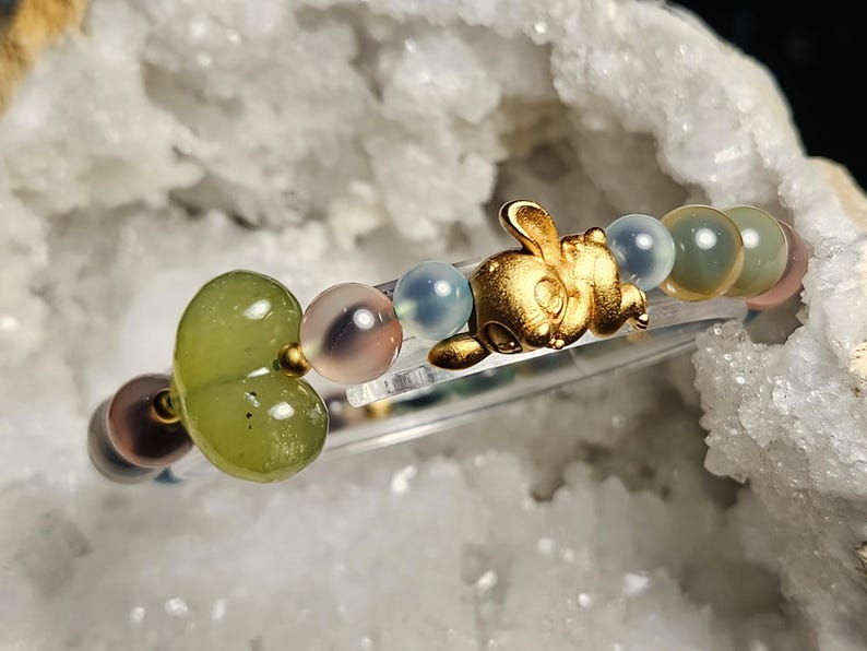 May include: A bracelet with a gold-coloured rabbit charm and assorted gemstone beads in green, pink, blue, and brown. The bracelet is strung on a clear elastic cord. The beads are round and smooth, with a polished finish.