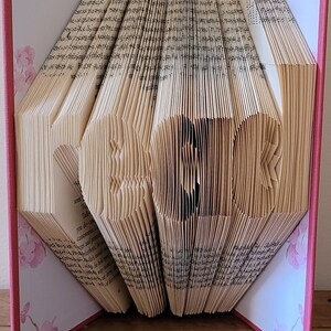 May include: A book sculpture with the word "BOOK" formed by folded pages. The book has a pink cover and floral patterned endpapers. It stands on a wooden surface. The pages are folded to create the letters.