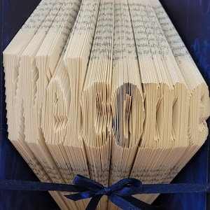 May include: A book art sculpture with the word "Welcome" folded into the pages. The book is bound in blue covers and has a blue ribbon tied around it.