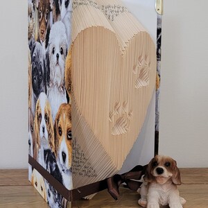 May include: A book shaped like a heart with a paw print design. The book is covered in fabric with a pattern of many different puppies. A small brown and white dog figurine is sitting in front of the book.