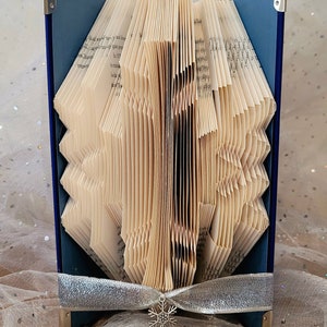 Op de afbeelding: Een blauwe boekkunst-sculptuur met een sneeuwvlokkenontwerp. Het boek is gevouwen tot een driedimensionale vorm en is versierd met een zilveren lint.