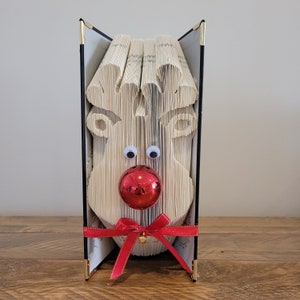 May include: A decorative book transformed into a reindeer face, with folded pages forming the shape. Features googly eyes, a red ornament nose, and a red ribbon with a small gold bell. The book has black covers with gold corner accents.