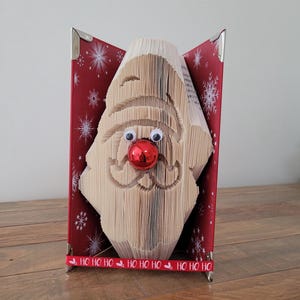 Könnte beinhalten: Handgefertigte Weihnachtsdekoration mit einem aus Buchseiten geformten Weihnachtsmann-Gesicht. Das Buch ist rot mit weißen Schneeflockenmustern und hat ein rotes Band mit dem Text "HO HO HO". Der Weihnachtsmann hat Wackelaugen und eine rote Nase.