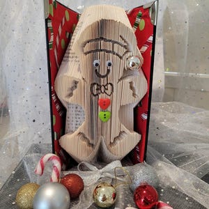 May include: A festive Christmas decoration made from a folded book, shaped into a gingerbread man. The gingerbread man has googly eyes, a bow tie, and button details. Candy canes and baubles are arranged around the book art.