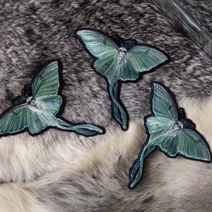 May include: Three green and white embroidered moth patches with a detailed design. The patches are on a grey and white fur background.