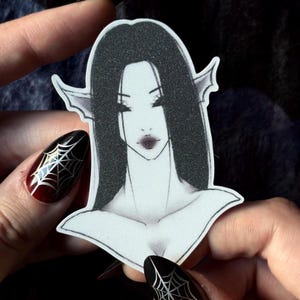 Peut inclure: Un autocollant représentant une illustration stylisée d'une femme aux longs cheveux noirs, aux oreilles pointues et au maquillage sombre. L'autocollant a une texture scintillante. Les ongles de l'image sont peints en noir et rouge avec un motif de toile d'araignée.