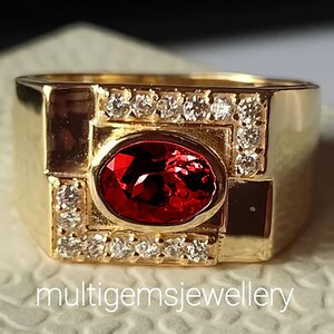 Puede incluir: Un anillo de oro con una piedra ovalada roja rodeada de diamantes más pequeños. El anillo tiene una banda cuadrada con un diseño geométrico.