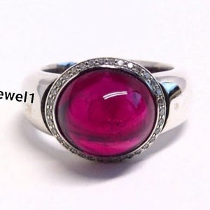 Puede incluir: Un anillo de plata con una gran piedra preciosa redonda de color rosa intenso en cabujón en el centro, rodeada por un borde fino de pequeñas piedras claras. El texto silvergemsjewel1 es visible en el lado izquierdo del anillo sobre fondo blanco.