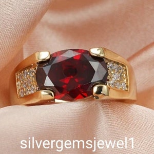 May include: A gold ring featuring a large, oval-shaped, red gemstone surrounded by smaller, clear gemstones.