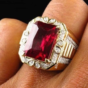Puede incluir: Un anillo dorado con una gran gema roja rectangular. El anillo está adornado con pequeñas gemas transparentes y filigranas decorativas. La banda tiene un diseño texturizado. El anillo está en el dedo de una persona.