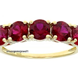 Puede incluir: Un anillo dorado con cinco piedras preciosas rojas ovaladas. Las piedras están dispuestas en una fila, creando una banda continua de color. La banda del anillo es lisa y pulida, con el texto "silvergemsjewel1" y "925" visible.
