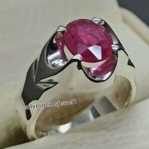 Puede incluir: Un anillo de plata con una gran gema de rubí de corte ovalado. El rubí es de color rojo intenso y está engastado en una banda de plata pulida. El anillo tiene un diseño clásico y elegante.